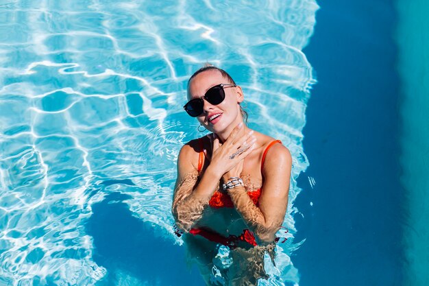 Porträt der ruhigen glücklichen Frau in der Sonnenbrille mit gebräunter Haut im blauen Schwimmbad am sonnigen Tag