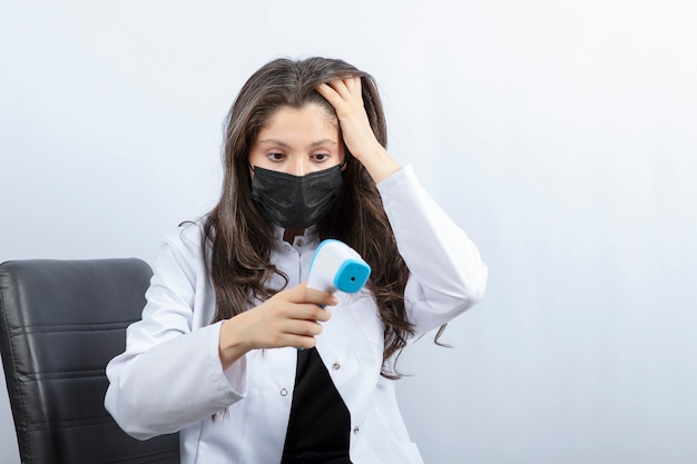 Porträt der Ärztin in medizinischer Maske und weißem Kittel mit Blick auf das Thermometer.