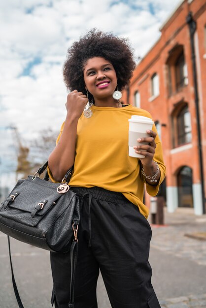 Porträt der schönen afroamerikanischen lateinischen Frau, die eine Tasse Kaffee draußen in der Straße geht und hält. Stadtkonzept.