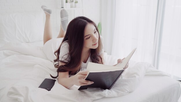 Porträt der schönen attraktiven Asiatin, die ein Buch beim Lügen auf dem Bett liest