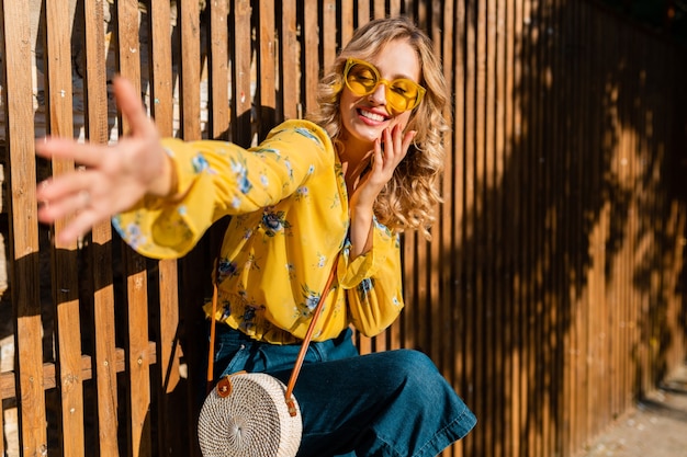 Porträt der schönen blonden emotionalen lachenden stilvollen lächelnden Frau in der gelben Bluse, die Sonnenbrille, Strohgeldbörse bali Art trägt