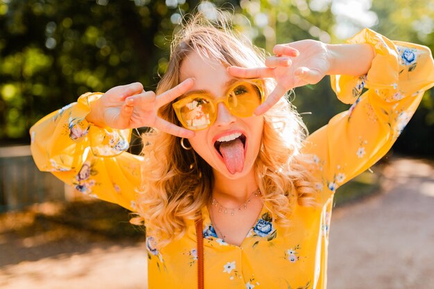 Porträt der schönen blonden stilvollen emotionalen Frau in der gelben Bluse, die Sonnenbrille, lustiger verrückter Gesichtsausdruck trägt