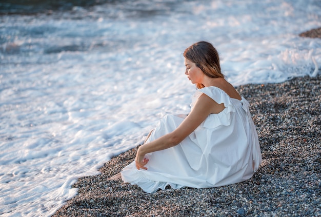 Porträt der schönen Frau an der Küste, die allein sitzt und im weißen Kleid denkt und traurig aussieht