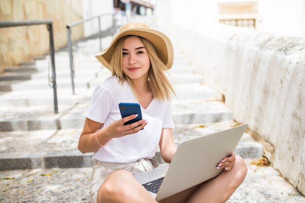 Porträt der schönen Frau, die Handy und Laptop hält, die auf der Sommerstadttreppe sitzen