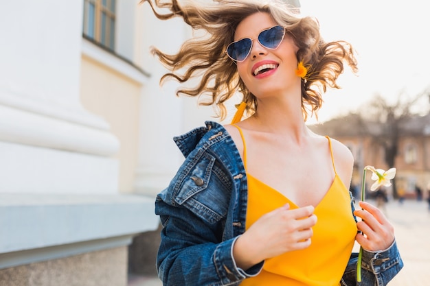 Porträt der schönen Frau, die lächelndes Haar winkt, stilvolle Kleidung, Jeansjacke und gelbes Oberteil tragend, Modetrend, Sommerstil, glückliche positive Stimmung, sonniger Tag, Sonnenaufgang, emotional, fröhlich