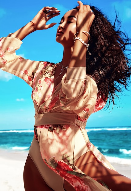 Porträt der schönen Frau mit dem dunklen langen Haar im beige Kleid, das am Sommerstrand aufwirft