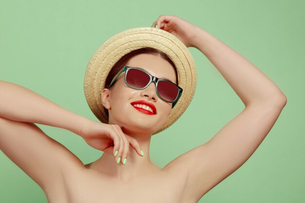 Porträt der schönen Frau mit hellem Make-up, Hut und Sonnenbrille auf grünem Studio