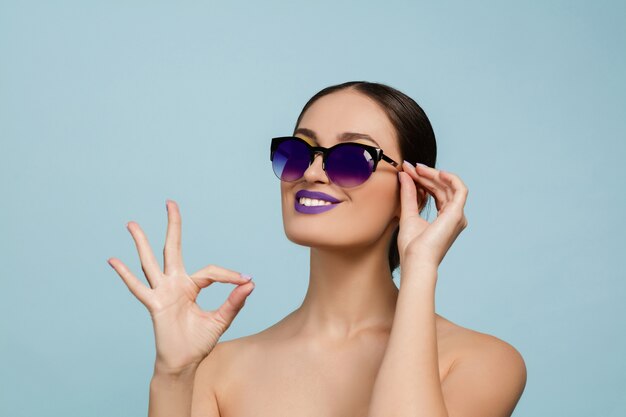 Porträt der schönen Frau mit hellem Make-up und Sonnenbrille auf blauem Studio
