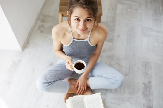 Porträt der schönen glücklichen Frau lächelnd, die Tasse Kaffee hält, der auf Boden mit Buch sitzt.