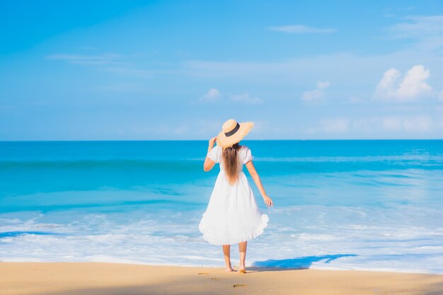 Porträt der schönen jungen asiatischen Frau, die um Strand mit weißen Wolken auf blauem Himmel im Reiseurlaub entspannt