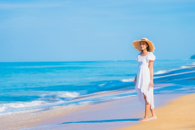 Porträt der schönen jungen asiatischen Frau, die um Strand mit weißen Wolken auf blauem Himmel im Reiseurlaub entspannt