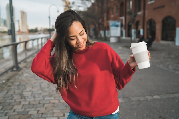 Porträt der schönen jungen Frau, die eine Tasse Kaffee draußen hält. Stadtkonzept.