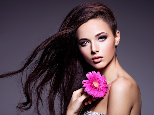 Porträt der schönen jungen Frau mit dem langen braunen Haar mit der rosa Blume, die über dunkler Wand aufwirft