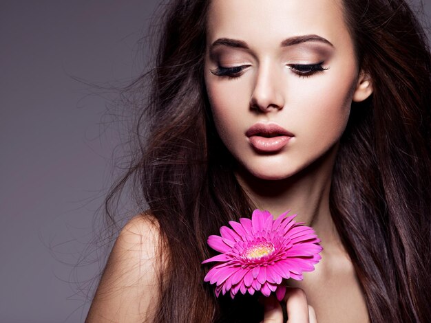 Porträt der schönen jungen Frau mit dem langen braunen Haar mit der rosa Blume, die über dunkler Wand aufwirft