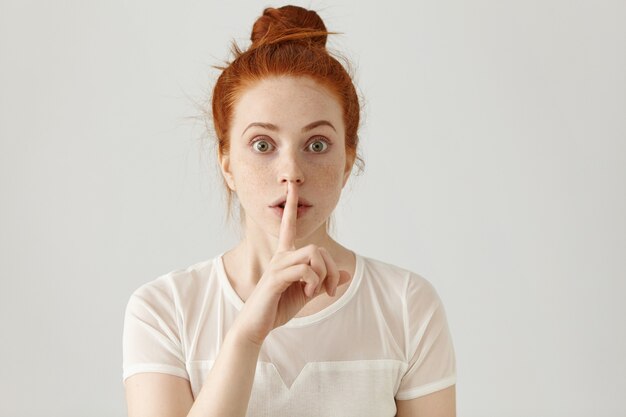 Porträt der schönen jungen kaukasischen Frau mit Ingwerhaarbrötchen, das Zeigefinger an den Lippen hält