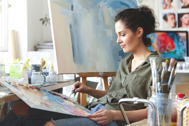 Porträt der schönen Künstlerin in Freizeitkleidung, die leuchtende Farben mischt, auf Staffelei zeichnet, während sie im Kunststudio sitzt. Brünette Malerin bei der Arbeit. Kreativität, Kunst, Malkonzept