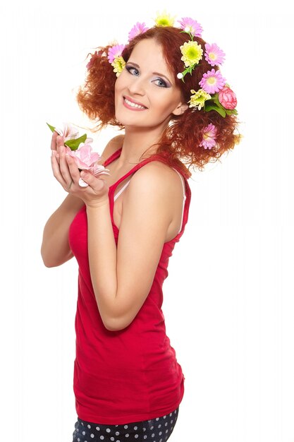 Porträt der schönen lächelnden rothaarigen Ingwerfrau im roten Stoff mit gelben rosa bunten Blumen im Haar lokalisiert auf weißen Halteblumen