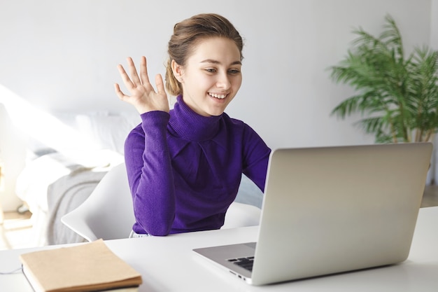 Porträt der schönen positiven jungen Frau, die Online-Kommunikation genießt, vor offenem Laptop sitzt, lächelt und Hand winkt, Hallo sagt
