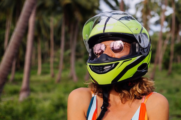 Porträt der schönen Reiterin im gelbgrünen Motorradhelm und im bunten hellen Sommerkleid im Dschungel auf tropischem Feld unter Palmen.