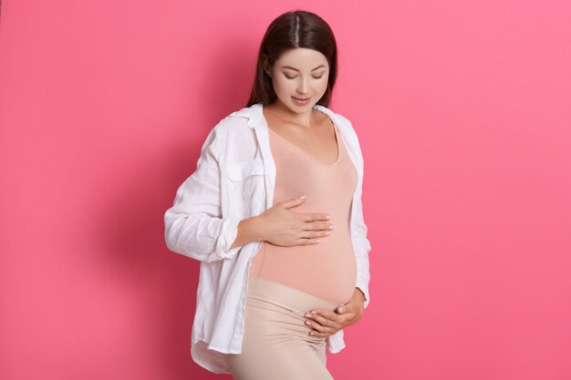 Porträt der schönen schwangeren Frau, die ihren Bauch hält und ihn mit großer Liebe betrachtet, während sie isoliert über rosigem Raum posiert, werdende Mutter, die stilvolle Kleidung trägt.