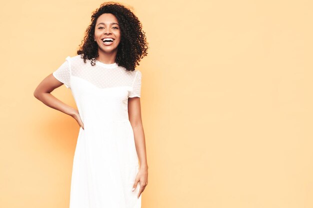 Porträt der schönen schwarzen Frau mit Afro-Lockenfrisur Lächelndes Modell in weißem Sommerkleid gekleidet Sexy sorglose Frau posiert in der Nähe der gelben Wand im Studio Gebräunt und fröhlich Isoliert