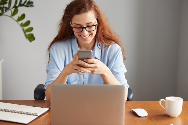 Porträt der schönen Sekretärin, die blaues Hemd und Brille trägt, lächelnd während der SMS