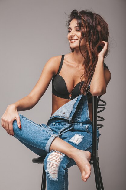 Porträt der sexy schönen Frau in den Jeans insgesamt. Nettes attraktives Hipster-Mädchen, das auf Stuhl sitzt. Model posiert im Studio