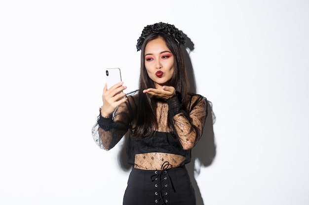 Porträt der stilvollen asiatischen Bloggerin mit gotischem Make-up und Halloween-Kostüm, das Luftkuss an der Handykamera sendet, Video aufzeichnet oder Videoanruf hat, der über weißem Hintergrund steht.