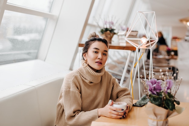 Porträt der stilvollen asiatischen Frau im beigen Pullover, der Tasse Kaffee hält