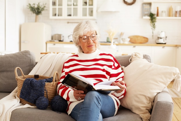 Porträt der stilvollen attraktiven Rentnerin in runden Brillen, die zu Hause mit gutem Buch entspannen. Freudige ältere Frau, die eine Brille trägt, Bestseller liest, bequem auf Sofa sitzt