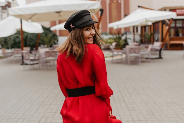 Porträt der stilvollen Dame in der schwarzen modischen Kappe, die ihren Kopf für Foto dreht. Mädchen im Satinkleid drehte ihr den Rücken zu