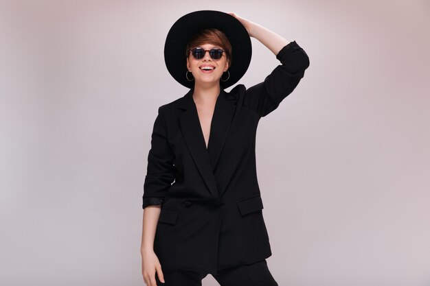 Porträt der stilvollen Dame in der Sonnenbrille und im Hut mit breiter Krempe. Kühle junge Frau in der schwarzen Jacke und in den Hosen stellt und lächelt auf lokalisiertem Hintergrund