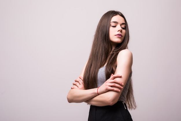 Porträt der stilvollen jungen hübschen Frau, die im grauen T-Shirt auf weißer Wand lächelt, lokalisierter, natürlicher Blick, langes braunes Haar, verschränkte Arme