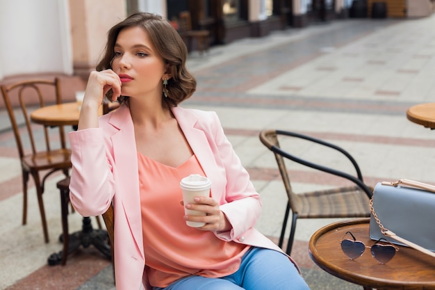 Porträt der stilvollen romantischen Frau, die im Café sitzt, das Kaffee trinkt, rosa Jacke und Bluse trägt, Farbtrends in der Kleidung, Frühlingssommermode, Accessoires Sonnenbrille und Tasche, denkend, schauend