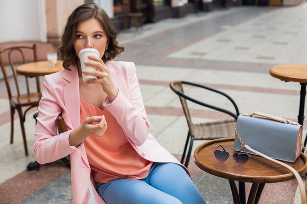 Porträt der stilvollen romantischen Frau, die im Café sitzt, das Kaffee trinkt, rosa Jacke und Bluse trägt, Farbtrends in der Kleidung, Frühlingssommermode, Zubehörsonnenbrille und Tasche