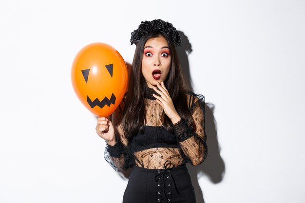 Porträt der überraschten asiatischen Frau im Halloween-Kostüm, als Hexe verkleidet, orange Ballon mit gruseligem Gesicht haltend.