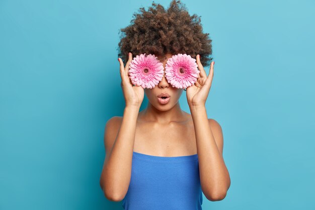 Porträt der überraschten jungen Frau mit lockigem Afro-Haar hält rosa Gerbera vor Augen hält Mund offen vom Wunder trägt Kleid posiert gegen blaue Wand