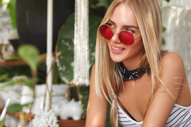 Kostenloses Foto porträt der verträumten entzückenden frau mit blondem haar leicht gebunden, trägt rote sonnenbrille