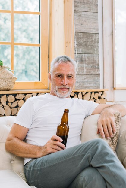 Porträt des älteren Mannes sitzend auf dem Sofa, welches die Kamera hält Bierflasche betrachtet