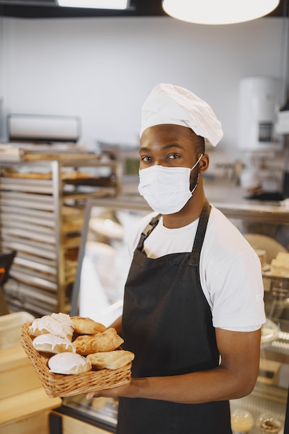 Porträt des afroamerikanischen Bäckers mit frischem Brot in der Bäckerei. Konditor, der kleines Gebäck hält.