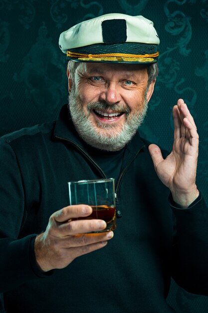 Porträt des alten Seemannsmannes als Kapitän im schwarzen Pullover und im Hut, der Cognac auf schwarzem Studio trinkt