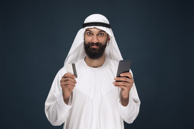 Porträt des arabischen saudischen Geschäftsmannes auf dunkelblauem Studiohintergrund. Mann mit Smartphone zum Bezahlen von Rechnungen, Online-Shopping oder Wetten. Konzept von Geschäft, Finanzen, Gesichtsausdruck, menschlichen Emotionen.