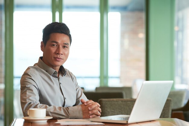 Porträt des asiatischen Geschäftsmannes von mittlerem Alter, der am Schreibtisch am Laptop sitzt