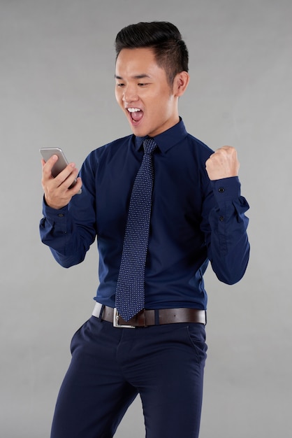 Porträt des asiatischen Kerls in der intelligenten Kleidung aufgeregt durch Smartphonnachrichten gegen grauen Hintergrund