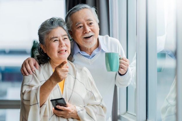 Porträt des asiatischen Seniorenpaares im Ruhestand, das lächelt und aus dem Fenster der Wohnung schaut, während es die Schulter der Frau hält