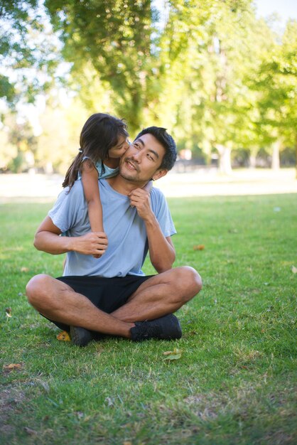 Porträt des asiatischen Vaters und der Tochter, die auf Gras im Park spielen. Glücklicher Mann, der auf dem Boden sitzt und ein kleines Mädchen, das seine Wange küsst und ihre Liebe zum Vater ausdrückt. Aktive Erholung und glückliches Kindheitskonzept