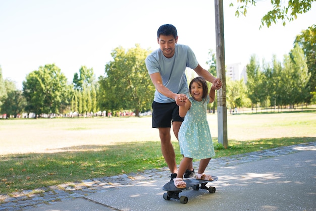 Porträt des asiatischen Vaters und des kleinen Mädchens, die zum Skateboard ausbilden. Lächelnder Mann, der auf der Gasse geht und die Hände der Töchter hält, während sie auf dem Skateboard fährt. Aktive Erholung, gesunder Lebensstil und Vaterschaftskonzept