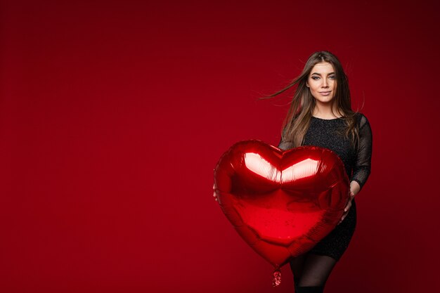 Porträt des atemberaubenden brünetten Mädchens im dunklen Cocktailkleid mit rotem Herzballon auf rotem Hintergrund. Konzept des Heiligen Valentinstags.