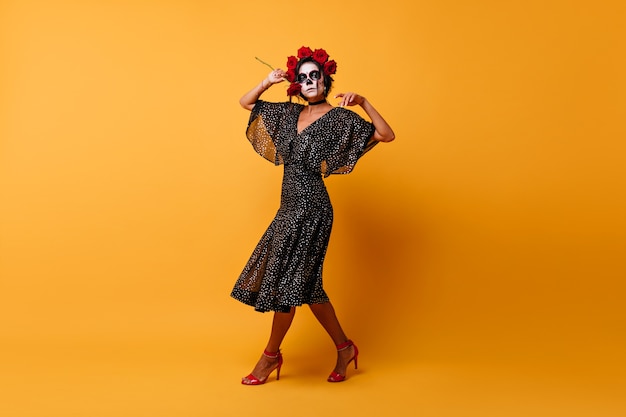 Porträt des atemberaubenden weiblichen Zombies in mexikanischer Kleidung in voller Länge. Inspiriertes schlankes Mädchen im Halloween-Kostüm, das auf orange Wand tanzt.