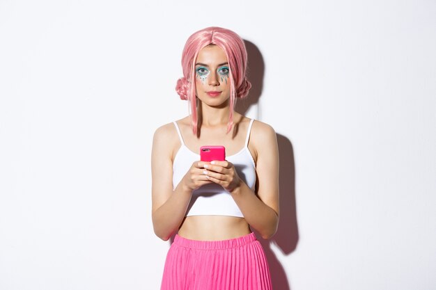 Porträt des attraktiven Partygirls in der rosa Perücke, mit dem hellen Make-up, das Smartphone hält und Kamera selbstbewusst betrachtet.